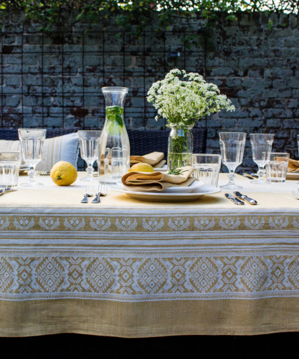 Yellow linen tablecloth with subtle motifs that reference Hungarian folk patterns. Draped over a table outside, set with glasses and tableware.