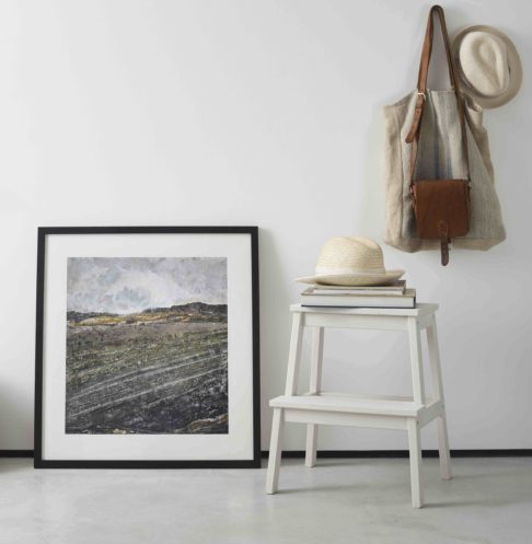 Northern Irish art print framed with a slim dark frame and leaned against a white wall next to a step stool with straw hats and bag on hook.