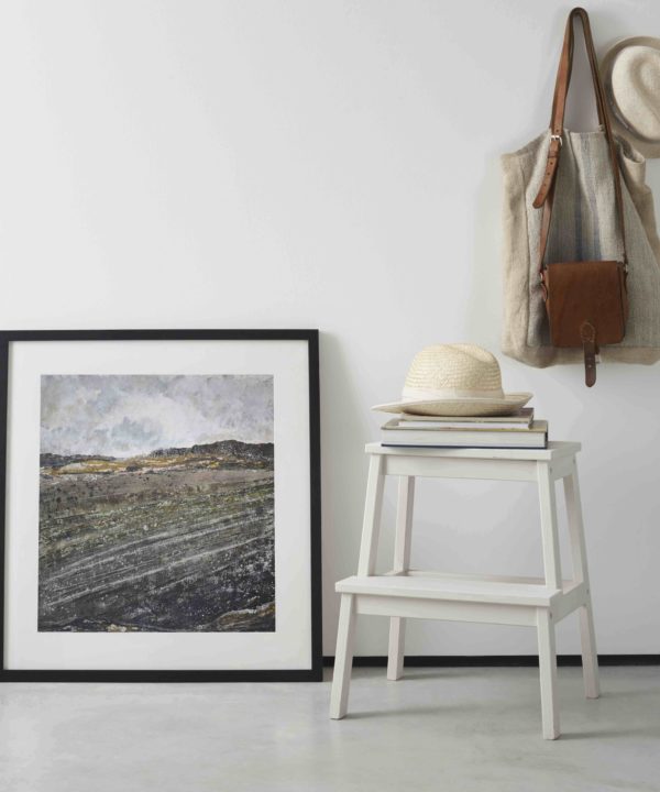 Northern Irish art print framed with a slim dark frame and leaned against a white wall next to a step stool with straw hats and bag on hook.