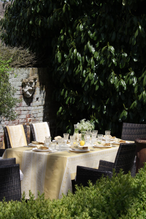 An outdoor entertaining table dressed with a soft yellow jacquard-woven tablecloth in a verdant garden setting.