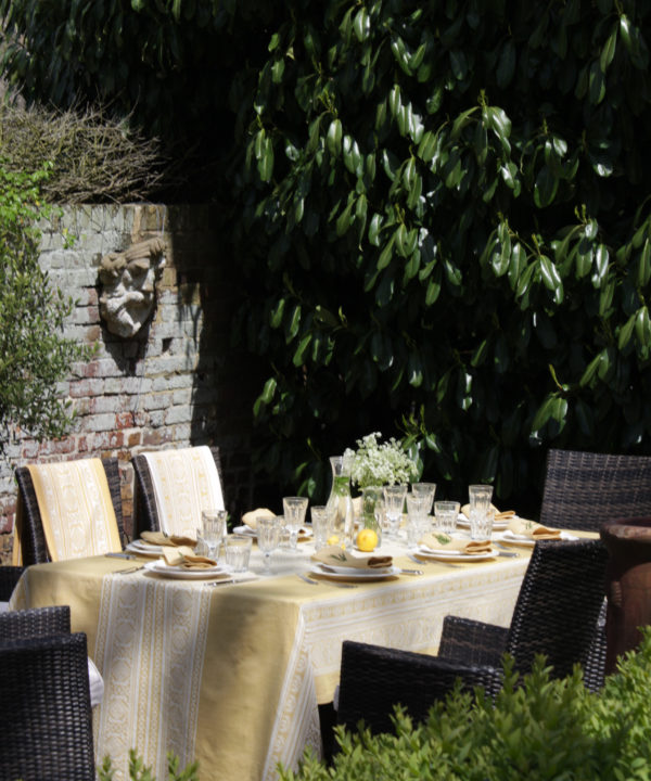 An outdoor entertaining table dressed with a soft yellow jacquard-woven tablecloth in a verdant garden setting.