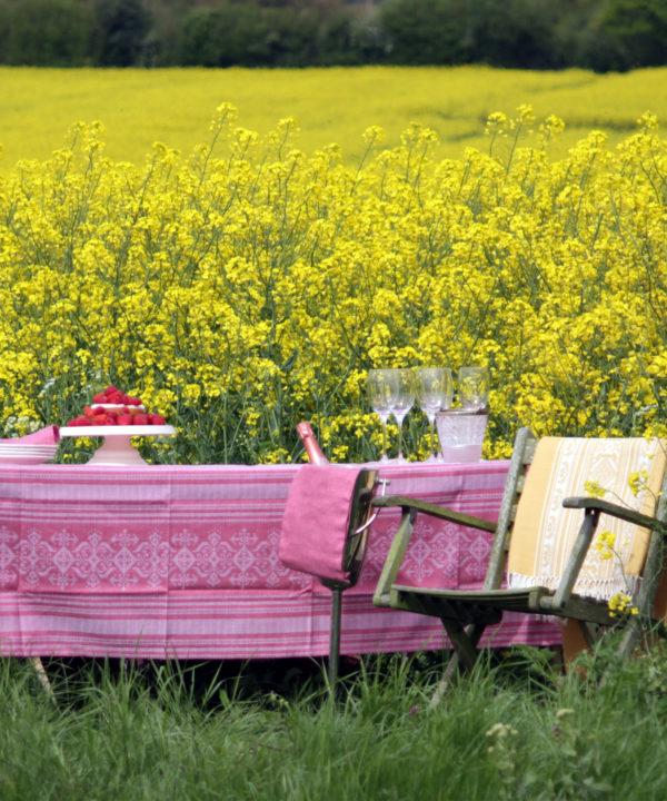 Pink linen tablecloth with subtle motifs that reference Hungarian folk patterns. Draped over a table in a field of yellow flowers.