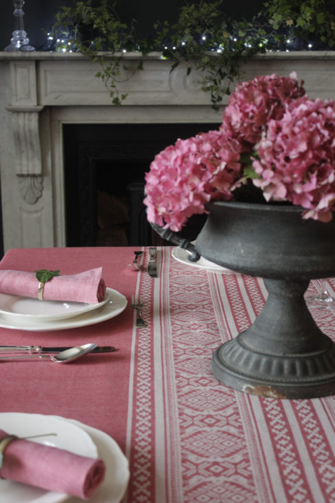 A deep pink Hungarian-folk-inspired tablecloth on a laid table with an urn filled with pink hydrangeas.