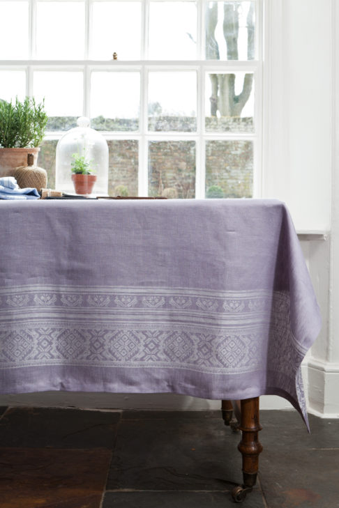 Purple tablecloth made from linen, with a Hungarian folk inspired jacquard weave, draped over a vintage mahogany dining table in front of a window.