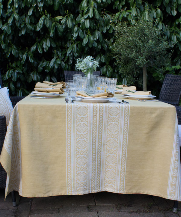 An elegant outdoor table set for supper with a soft yellow tablecloth depicting jacquard-woven, Hungarian folk patterns.