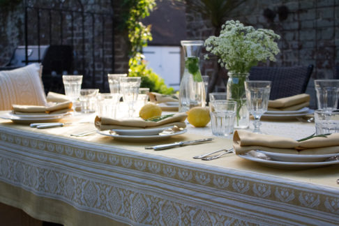 An outdoor table setting on a summer evening laid up with flowers, crockery, glassware, and a yellow Hungarian-folk-inspired tablecloth.