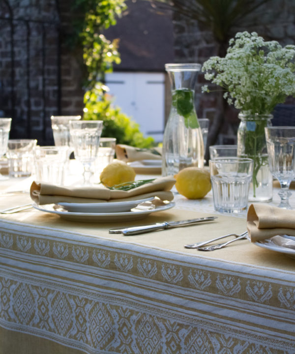 An outdoor table setting on a summer evening laid up with flowers, crockery, glassware, and a yellow Hungarian-folk-inspired tablecloth.