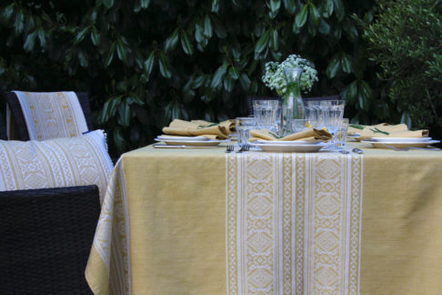 Detail of an outdoor table set for supper with a soft yellow tablecloth depicting jacquard-woven, Hungarian folk patterns.