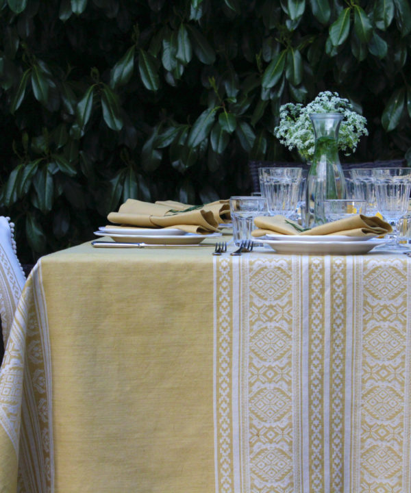 Detail of an outdoor table set for supper with a soft yellow tablecloth depicting jacquard-woven, Hungarian folk patterns.