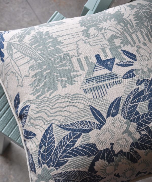 A hand-printed linen cushion in soft blues, depicting an Arts and Crafts house in a garden filled with rhododendrons.