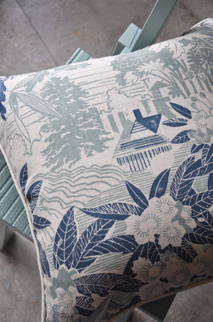 A hand-printed linen cushion in soft blues, depicting an Arts and Crafts house in a garden filled with rhododendrons.