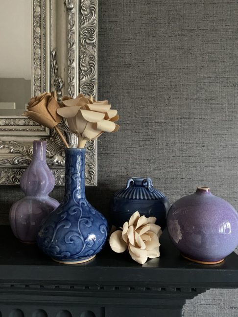 A selection of handmade Thai pottery displayed on a dark mantelpiece shelf against a silver mirror and grey textured wallpaper.