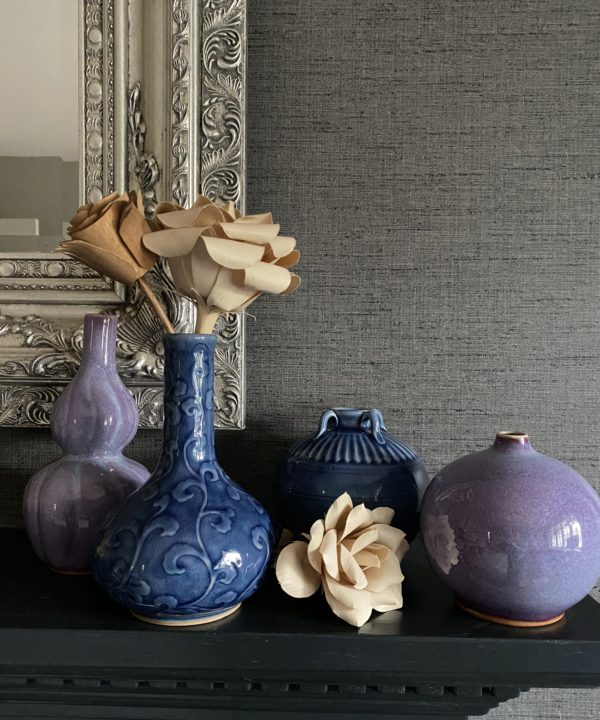 A selection of handmade Thai pottery displayed on a dark mantelpiece shelf against a silver mirror and grey textured wallpaper.