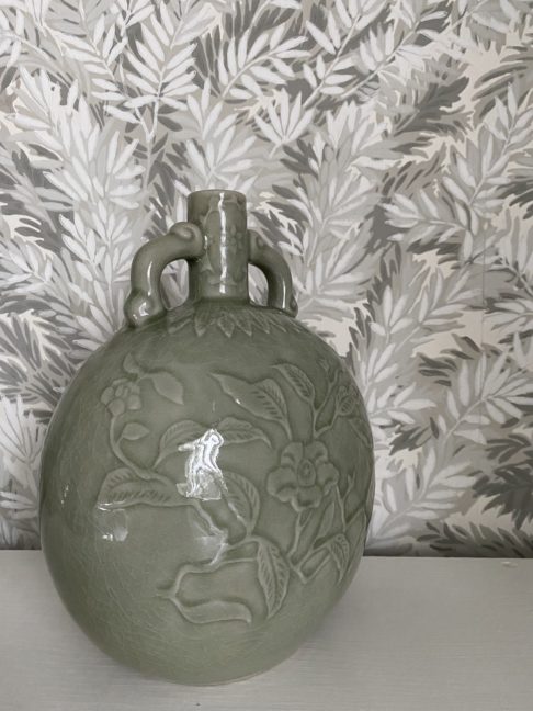 An ovoid, celadon pottery pot handmade in Thailand displayed against soft grey-green yew tree wallpaper.