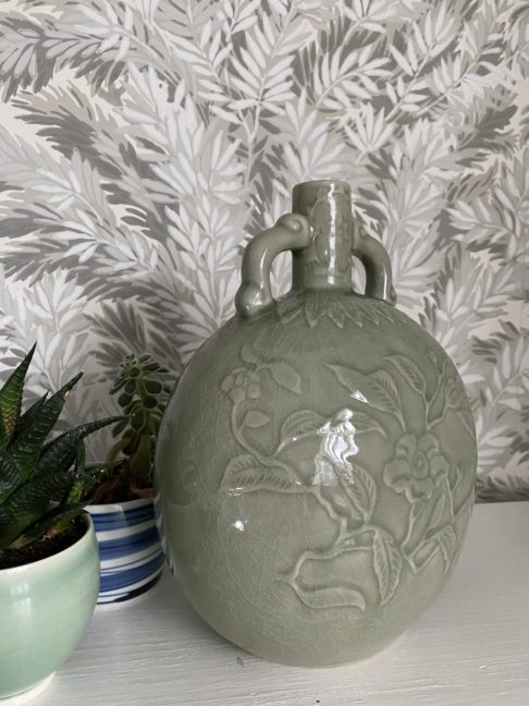 An elegant, celadon pottery pot with hand-decorated detailing photographed against a Cole & Son wallpaper depicting yew tree foliage in soft grey-greens.