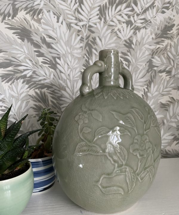 An elegant, celadon pottery pot with hand-decorated detailing photographed against a Cole & Son wallpaper depicting yew tree foliage in soft grey-greens.