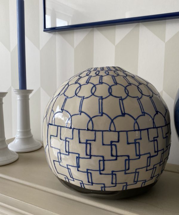 Geometric pottery pot in blue and cream on a beige bookshelf with white candlesticks behind.