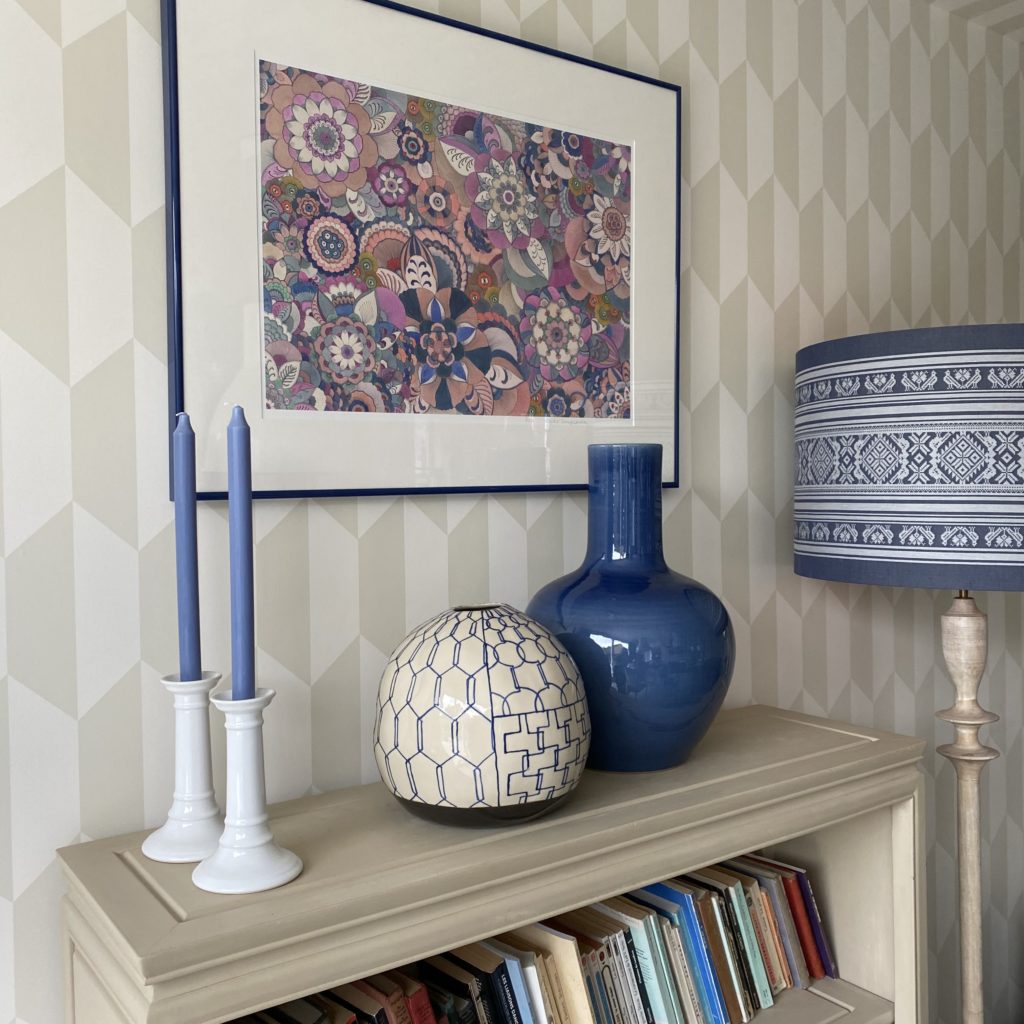 A still life display of Asian ceramics on a beige bookshelf with candlesticks and a pink floral Japanese artwork on the wall.