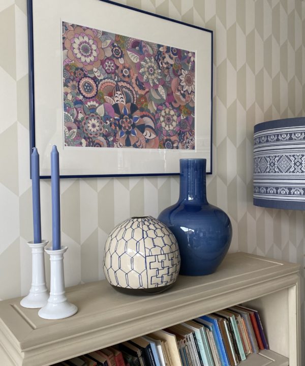 A still life display of Asian ceramics on a beige bookshelf with candlesticks and a pink floral Japanese artwork on the wall.