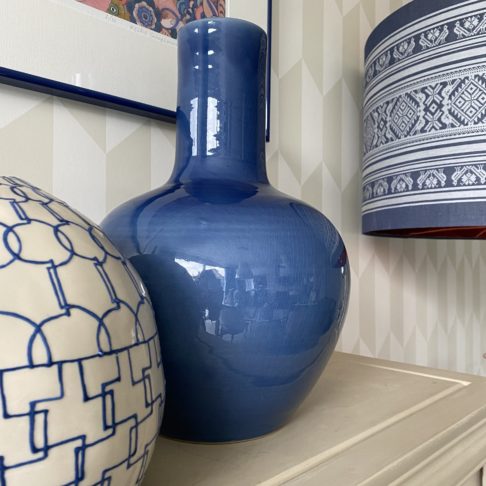 A striking, large, decorative blue pot on a pale beige surface with other ceramics and a patterned blue and white lampshade in the background.