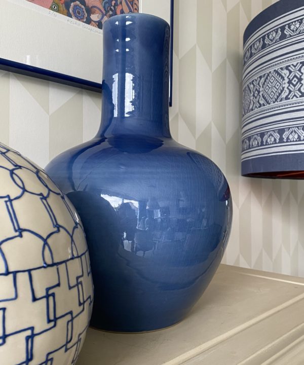 A striking, large, decorative blue pot on a pale beige surface with other ceramics and a patterned blue and white lampshade in the background.