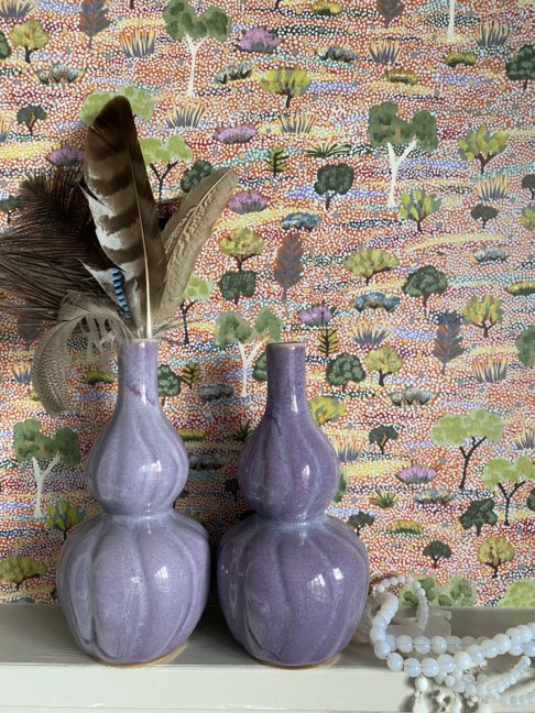 Purple ceramic pot, handmade in Thailand, double-gourd-shaped and shown holding feathers against a pretty, pastel, Aboriginal art wallpaper.