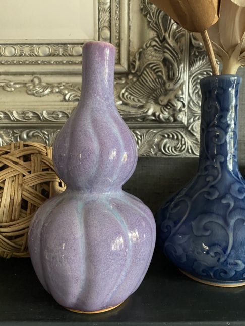 A shapely, purple ceramic pot handmade in Thailand sat a dark shelf with a silver mirror and other ceramics behind.