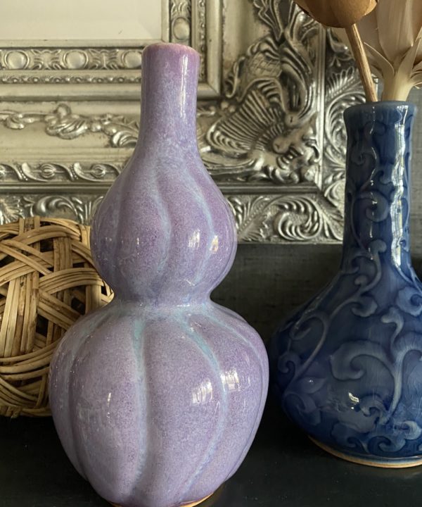 A shapely, purple ceramic pot handmade in Thailand sat a dark shelf with a silver mirror and other ceramics behind.