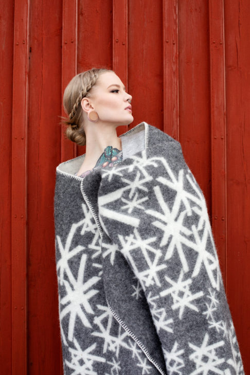 Grey blanket throw with Nordic bindrunes on a model against a red, wooden panelled shed.