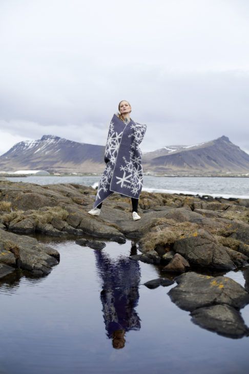 An Icelandic runes motif blanket in grey styled on a model in the Icelandic countryside with sea and mountains as a backdrop.