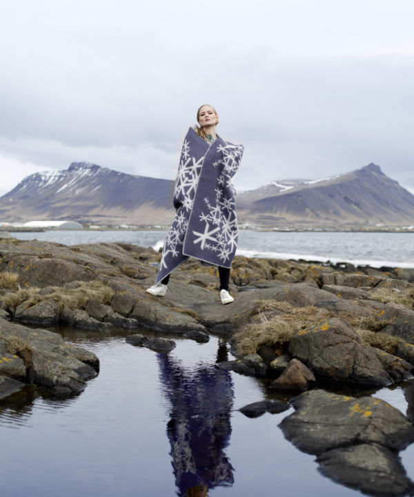 An Icelandic runes motif blanket in grey styled on a model in the Icelandic countryside with sea and mountains as a backdrop.