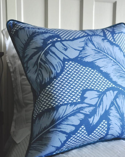 A blue tropical banana leaf print cushion on a bed.