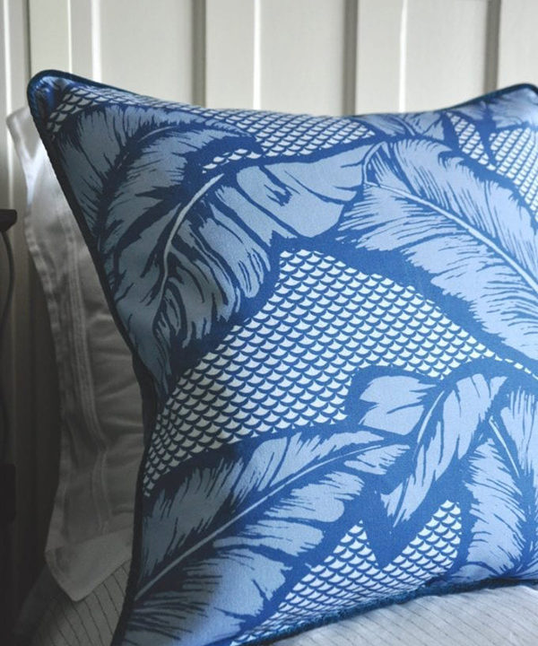 A blue tropical banana leaf print cushion on a bed.