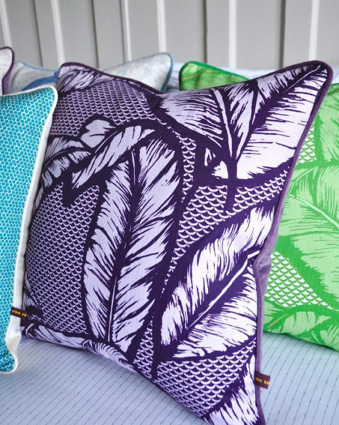 A deep purple Banana leaf print cushion with purple velvet back on a bed with other colourful cushions.