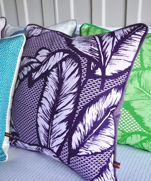 A deep purple Banana leaf print cushion with purple velvet back on a bed with other colourful cushions.