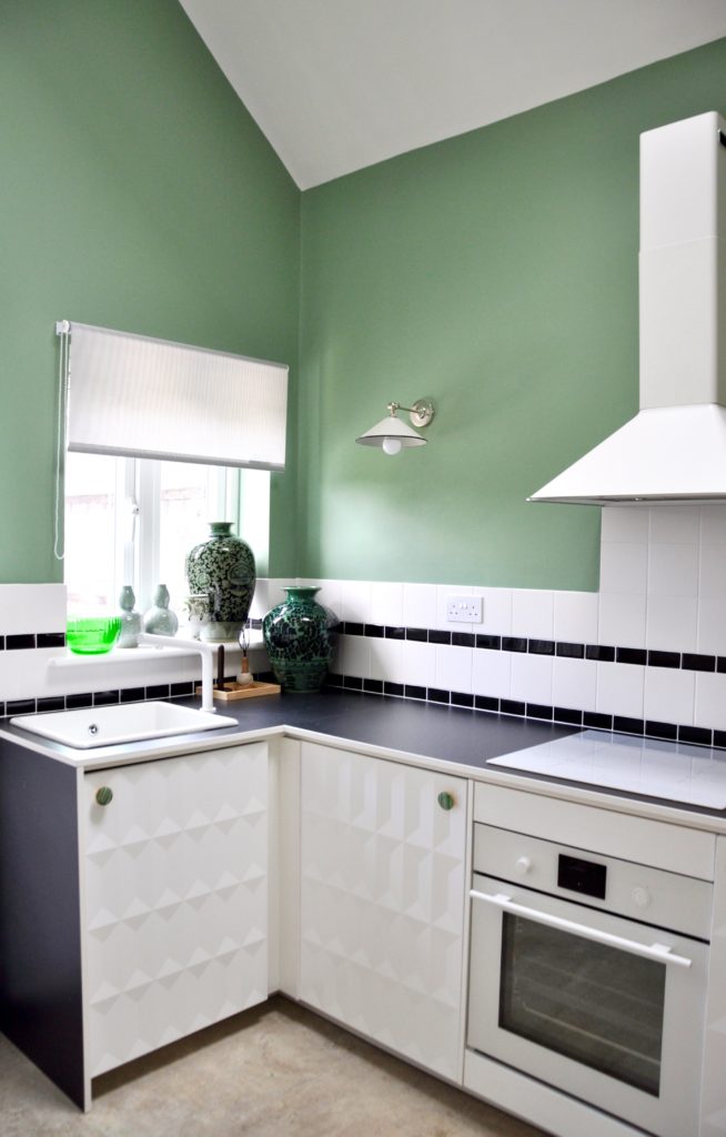 A modern, white, compact kitchen with black worktop, sage-green walls and monochrome, tiled splashback.