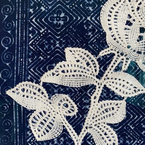 Textile art close up shot of a bobbin lace rose framed against indigo textiles.