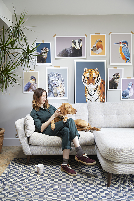 Digital artist, Talia Giles, on a sofa with her dog with a selection of her animal portraits on the wall behind.