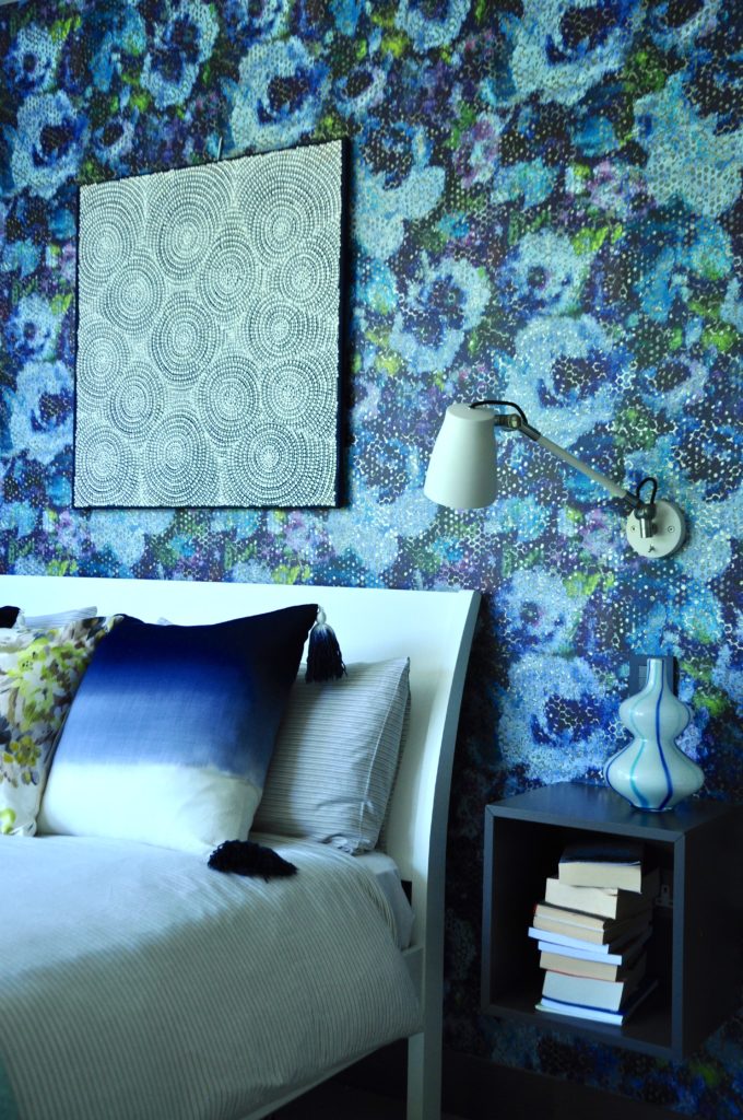 A modern, white ed against a blue floral Designers Guild wallpaper with anglepoise reading lamp and wall-hung bedside unit.