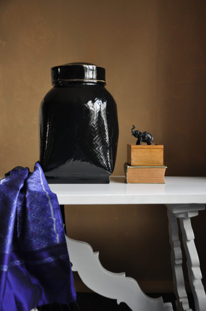 A gloss black lidded urn made from woven grasses in Thailand is displayed on a white Italian table with vibrant purple silk and a tiny, elephant model.