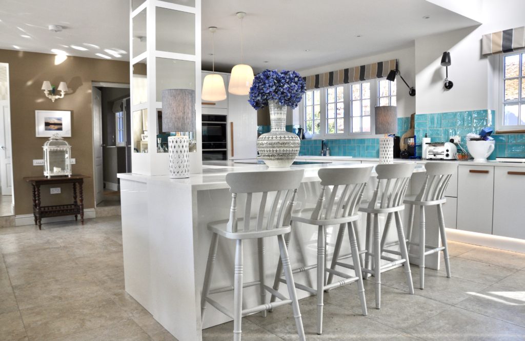 A large white island replete with a mirrored pillar in an eclectic white kitchen with a giant Thai ceramic pot, a turquoise-tiled splashback and vintage wooden furniture.
