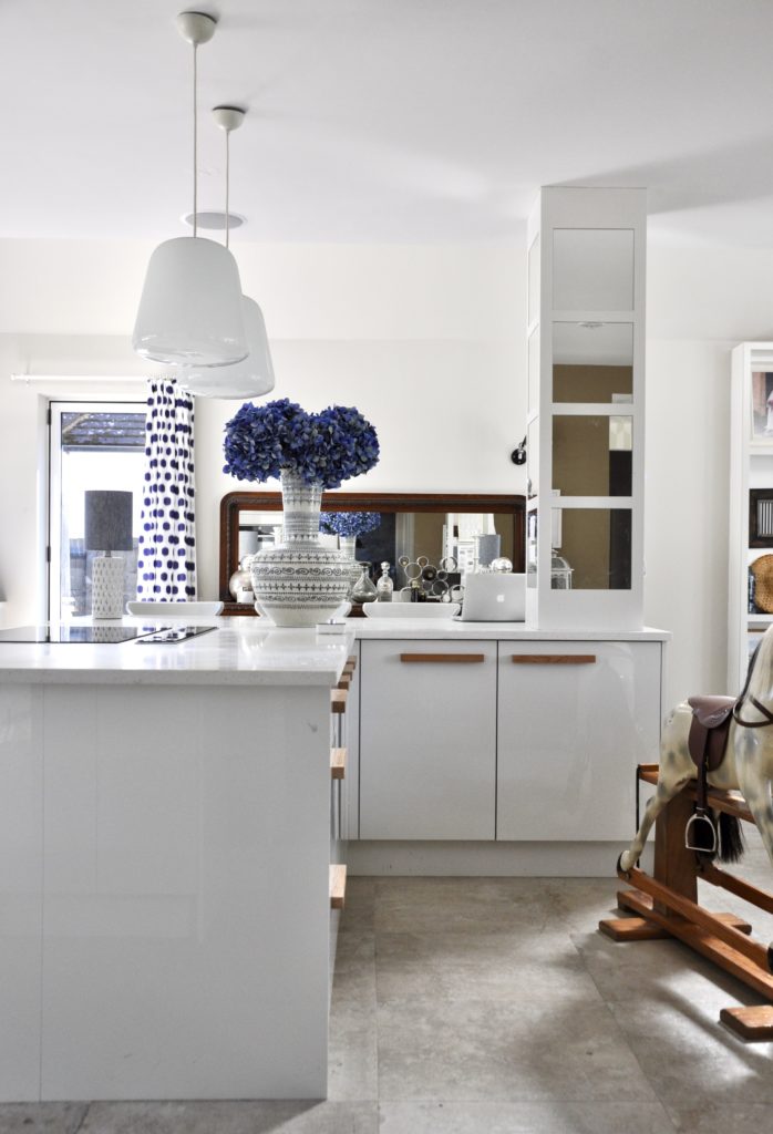 An L-shaped island in an eclectic white kitchen with a giant Thai ceramic pot on display, a polka dot blue and white curtain and a wooden sideboard.