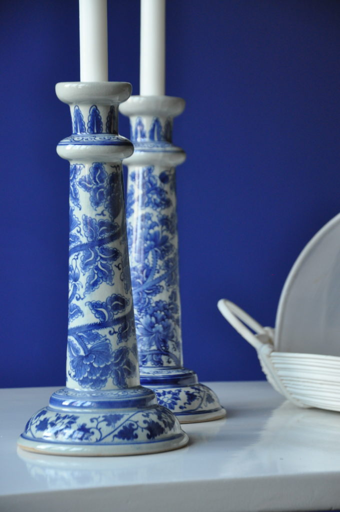 A pair of hand-painted Thai candlesticks in blue and white against a blue backdrop on a white table.