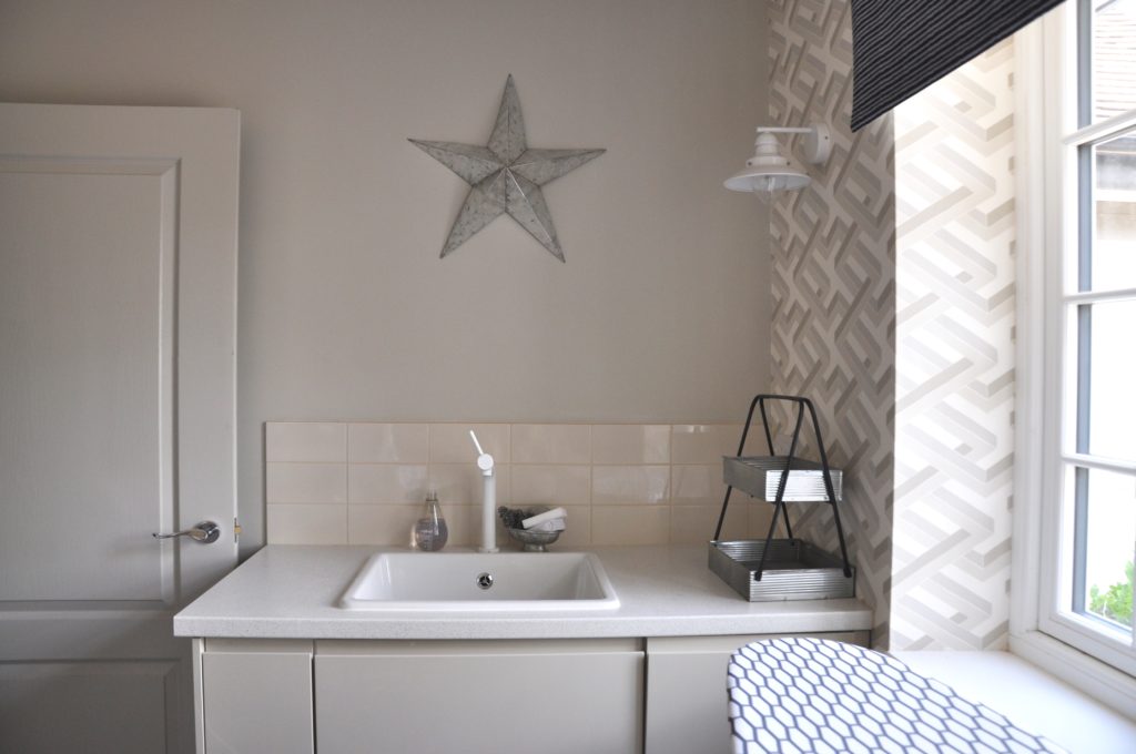 A contemporary laundry room with geometric wallpaper from Cole & Son, neutral tiles and units and a metal star on the wall.