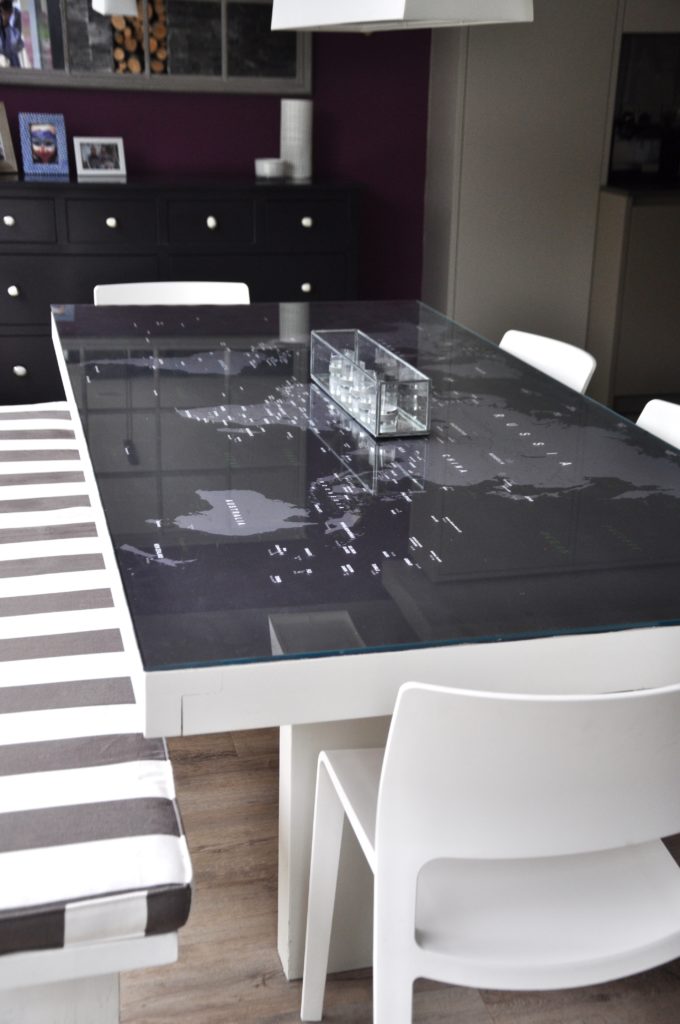 A glass table top with a grey world map set beneath it alongside a striped cushion on a bench.