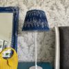 Indigo blue lampshades for table lamps displayed here on a simple, white candlestick lamp base on a teal sideboard with a blue decorative mirror and yellow wicker hand bag.