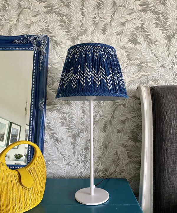 Indigo blue lampshades for table lamps displayed here on a simple, white candlestick lamp base on a teal sideboard with a blue decorative mirror and yellow wicker hand bag.