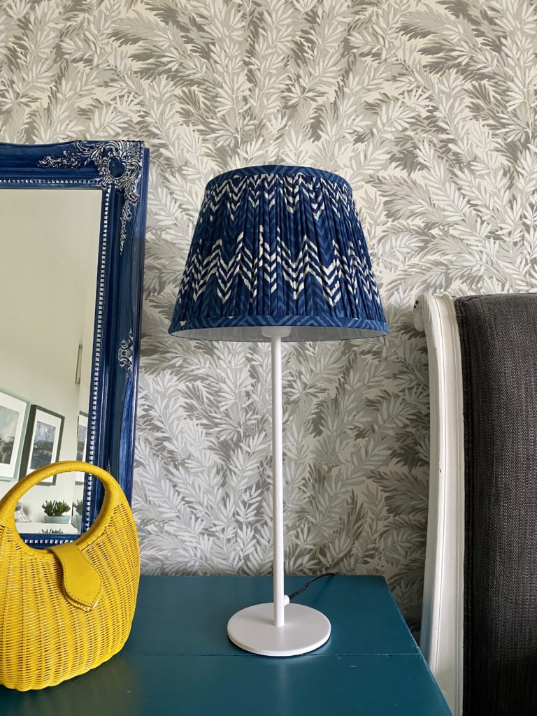 Indigo blue lampshades for table lamps displayed here on a simple, white candlestick lamp base on a teal sideboard with a blue decorative mirror and yellow wicker hand bag.