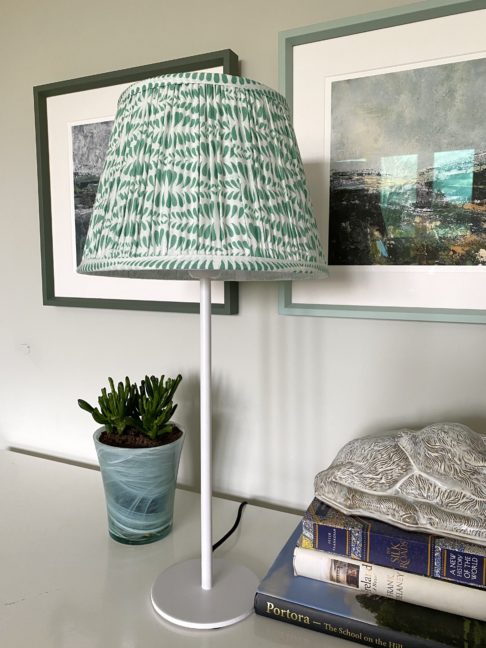 A mint-coloured, block-print lampshade in gathered cotton on a desk with Irish landscape prints in the background.