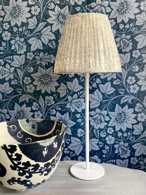 A vintage silk lampshade on a white candlestick base on a table with black and white bowl and floral folk wallpaper in dark blue behind.